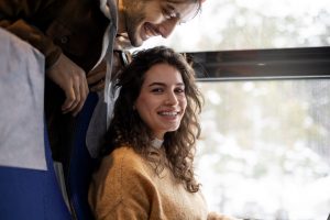 Mulher sorrindo, sentada na janela do ônibus, enquanto amigo está me pé atrás do banco, conversando sobre os motivos para viajar de ônibus.