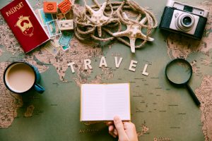 Jovem segurando um caderno com seu roteiro de viagens, em cima de uma mesa com um mapa, estrelas do mar, corda, papéis, passaporte, câmera fotográfica e uma xícara de café.