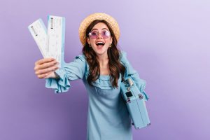 Menina jovem com mala azul, usando um chapéu e óculos, mostra animada suas passagens de ônibus que conseguiu efetuar uma troca e devolução.