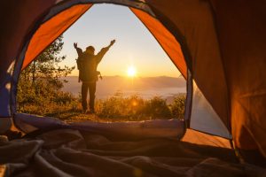 Homem em frente à barraca do seu acampamento em São Paulo, apreciando o nascer do sol no horizonte.