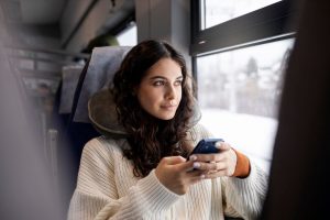 Mulher jovem sentada em uma poltrona do ônibus, olhando pela janela, enquanto pesquisa por que viajar de ônibus é a melhor alternativa.